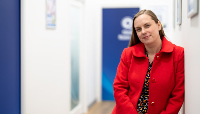 Elise Apolloni PSA Elise Apolloni, a community spirited pharmacist, standing in a pharmacy hallway wearing a red coat